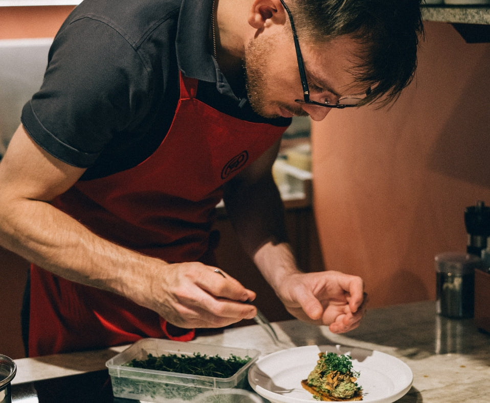 Kock med rött förkläde dekorerar en rätt med örter i restaurangkök – fokus på hantverk och julstämning.