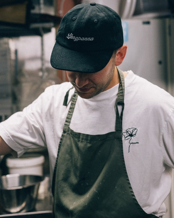 Kock med svart keps med brodyr och grönt förkläde arbetar i kök.