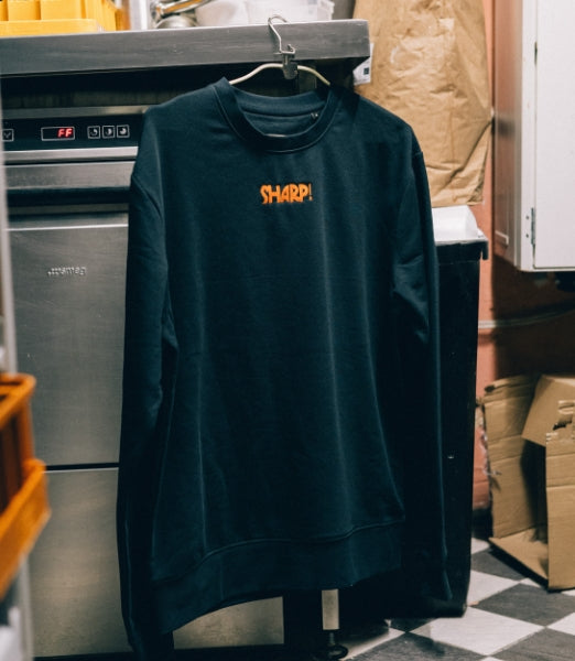 Svart tröja med orange brodyrtext “SHARP!” hänger på galge i kök.