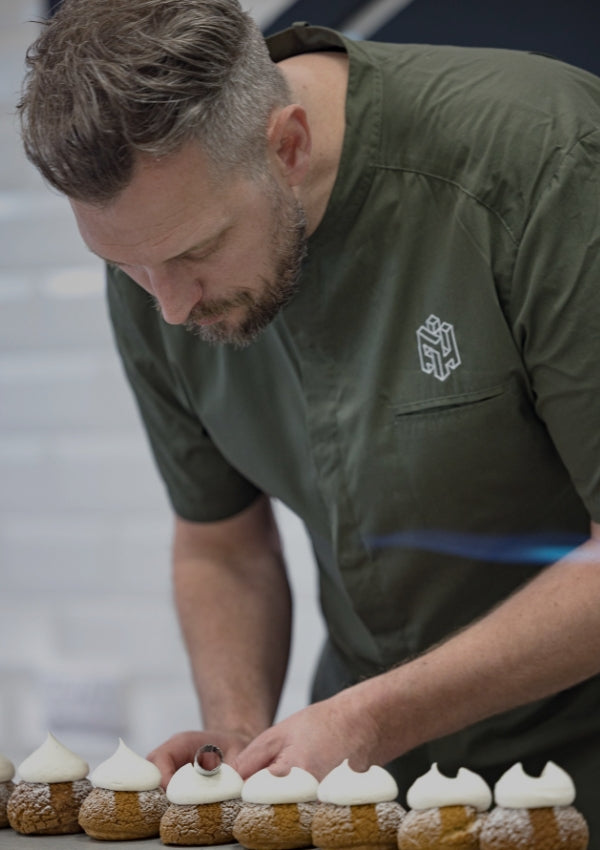 En bagare i grön kockrock dekorerar bakverk med maräng i ett modernt bageri.