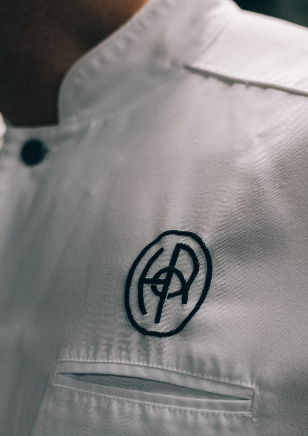 Närbild på en vit kockrock med ett broderat monogram i svart tråd på bröstet.