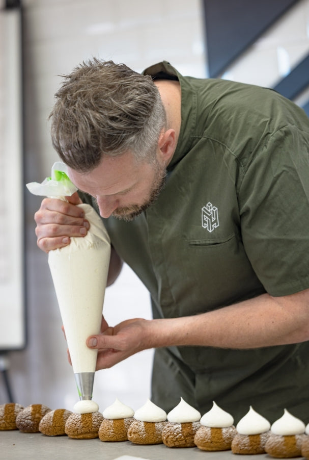 Konditor i grön kockrock spritsar grädde på en rad bakverk i ett bageri.