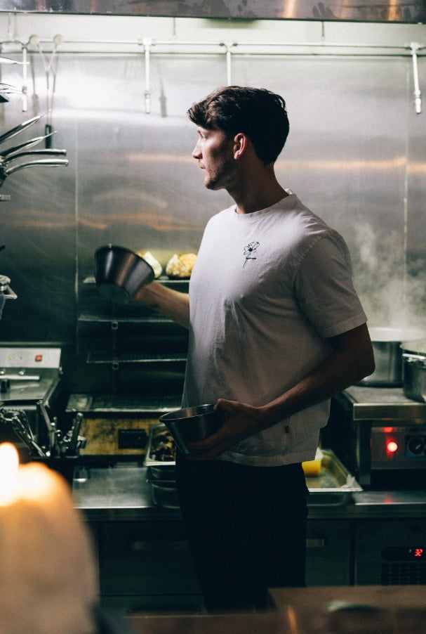 Kock i vitt t-shirt med broderad blomma arbetar i ett restaurangkök.