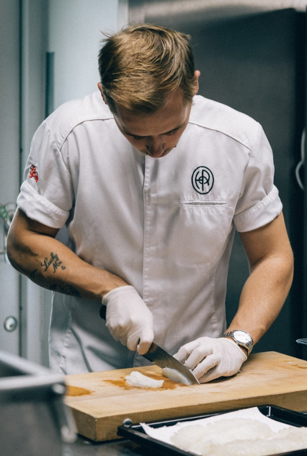 Kock i vit kockrock skär fisk på en skärbräda i kök.