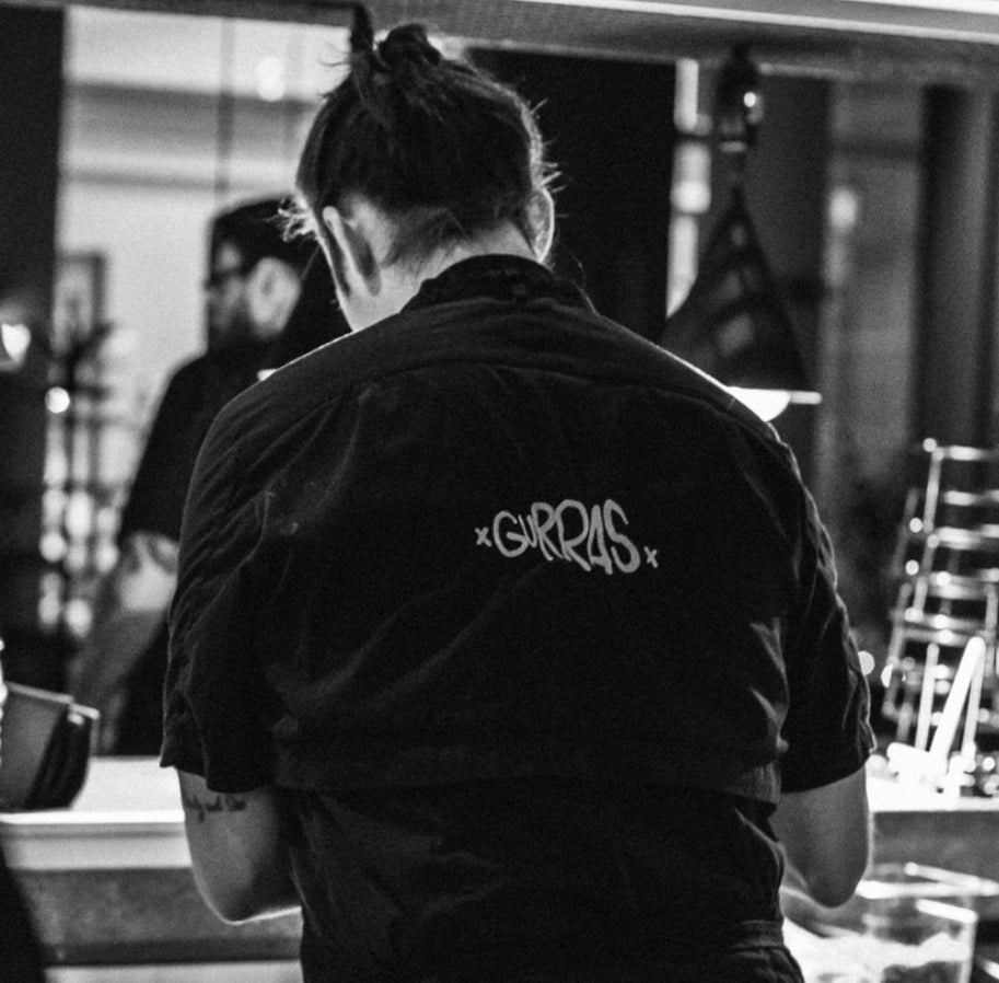 En kock i svart kockrock med texten "Gurras" på ryggen arbetar i ett restaurangkök.