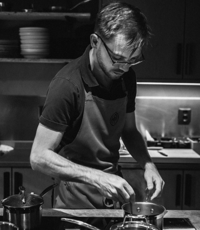 Kock i förkläde rör i kastrull på spis i svartvitt foto – professionell matlagning i köksmiljö.