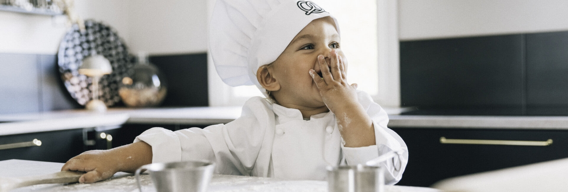 Litet barn i vit kockrock och kockmössa skrattar och leker i köket med mjöl på händerna. Kockkläder i barnstorlek från Segers.