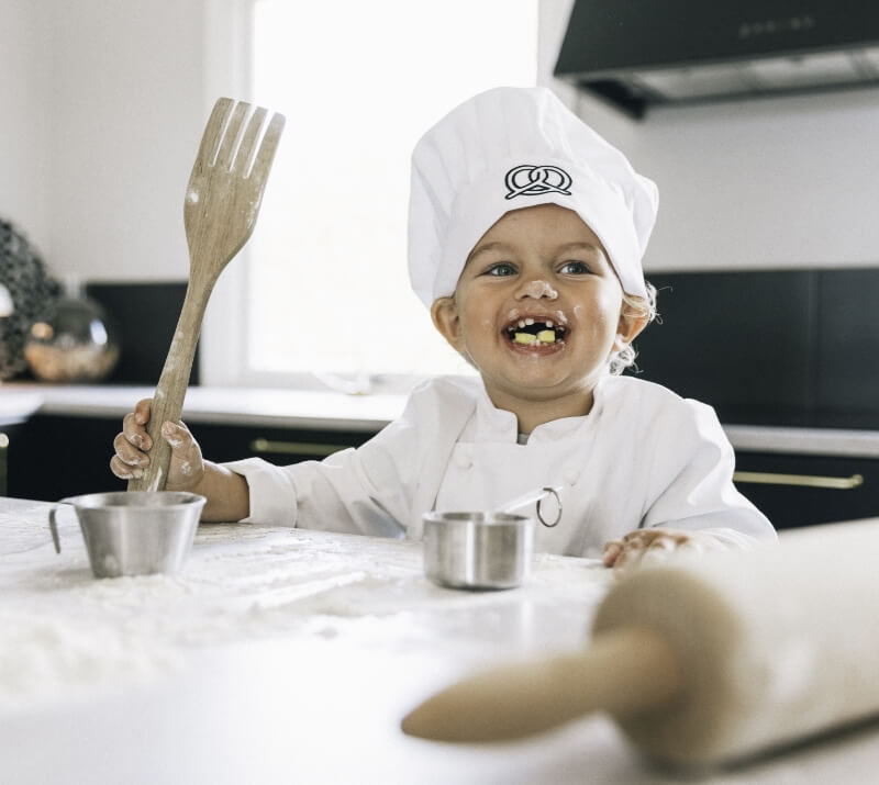 Leende barn i vit kockrock och kockmössa sitter i köket med en stor träslev. Kockkläder i barnstorlek från Segers.