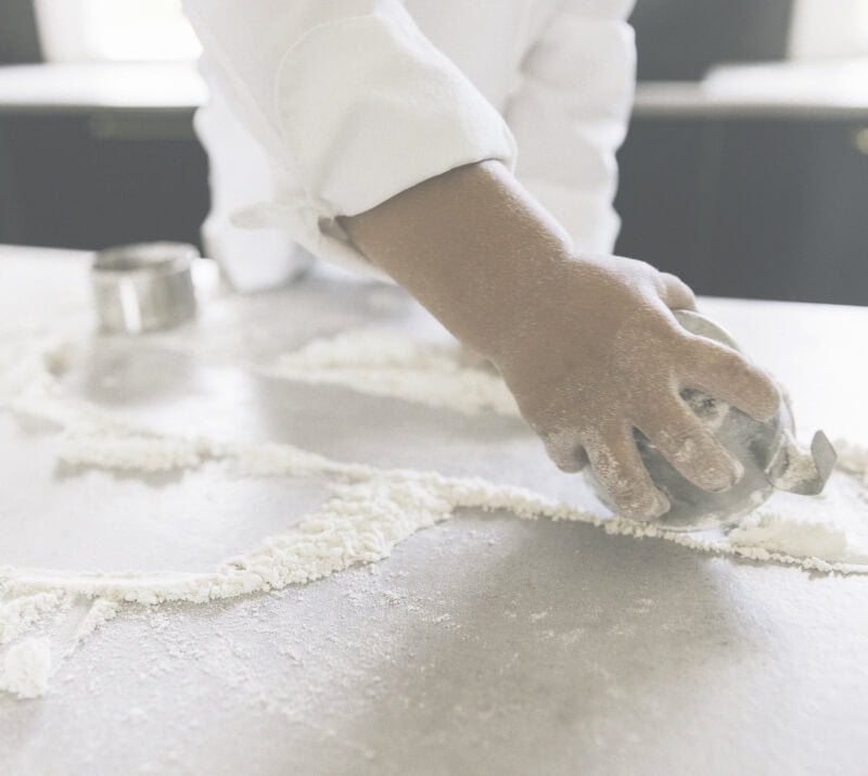 Närbild på barns hand i kockkläder som arbetar med mjöl på en köksbänk. Bakning med kockkläder barn.