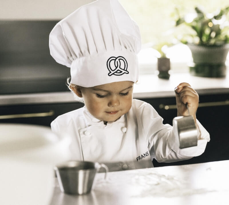 Barn i kockkläder håller i ett mått och bakar i köket. Vit kockrock och kockmössa med logotyp.