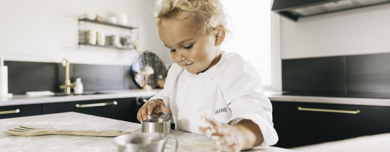 Barn i vit kockrock fokuserar på att mäta upp ingredienser vid bakning i köket. Kockkläder barn från Segers.