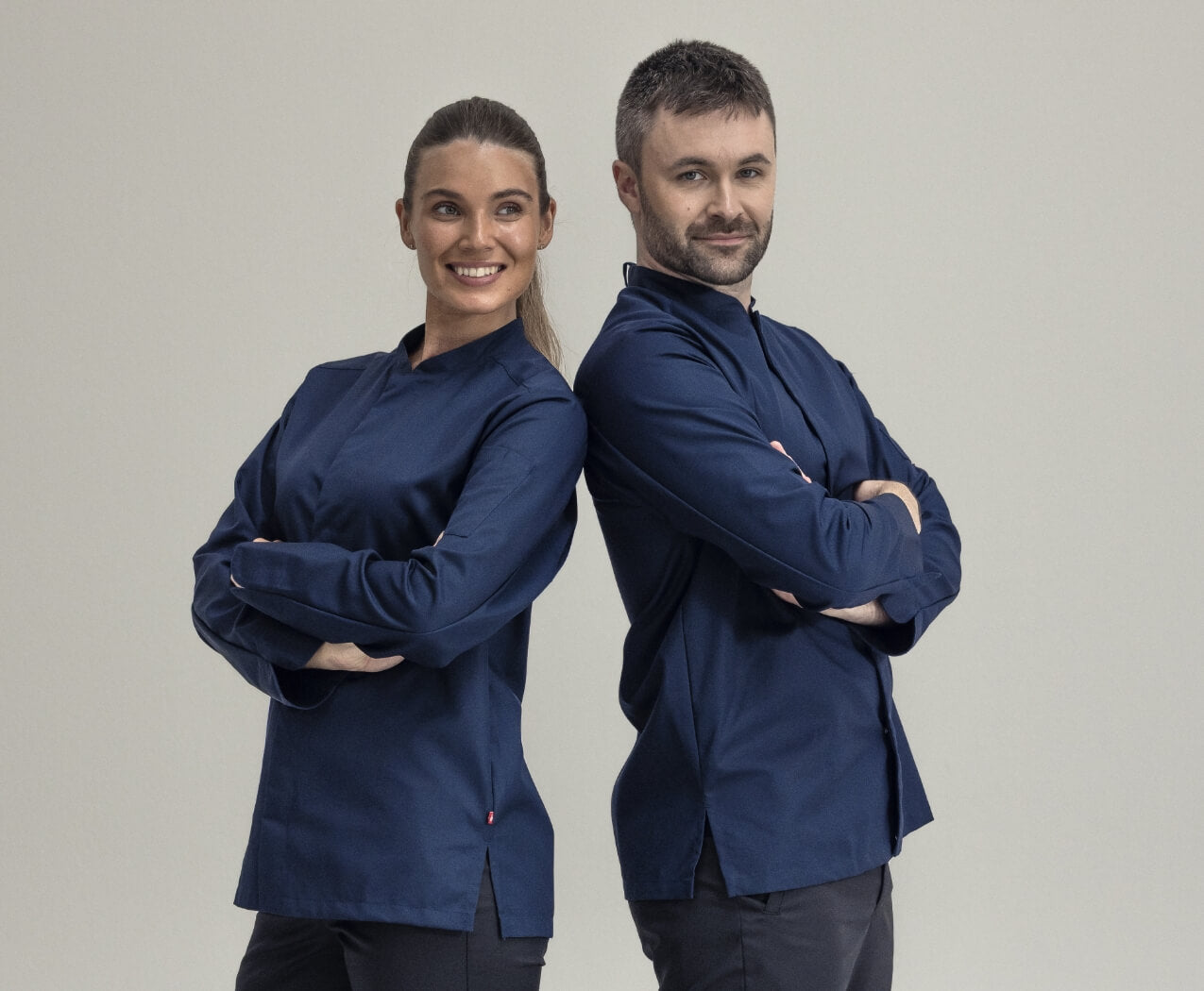 Två personer står rygg mot rygg med armarna i kors och ler. De bär mörkblå långärmade arbetsjackor och svarta byxor mot en ljus bakgrund.